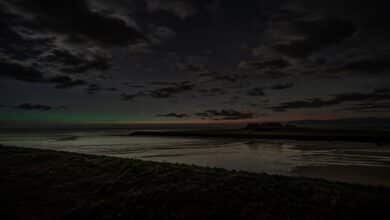 KI-Illustration einer Nordseeküste bei Nacht mit niedrigem grünem Polarlichtbogen am Horizont zwischen Wolken, kein reales Foto.