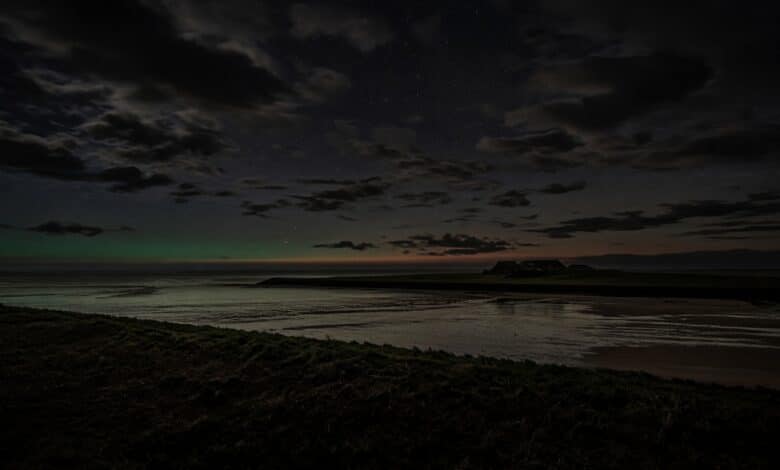 KI-Illustration einer Nordseeküste bei Nacht mit niedrigem grünem Polarlichtbogen am Horizont zwischen Wolken, kein reales Foto.